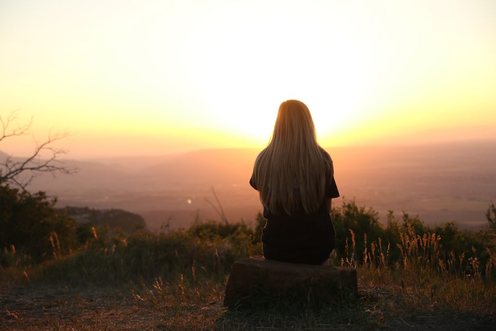 Woman sat on rock on a mountain top in her thoughts watching the sun set in the horizon. This woman is developing her positive mindset
