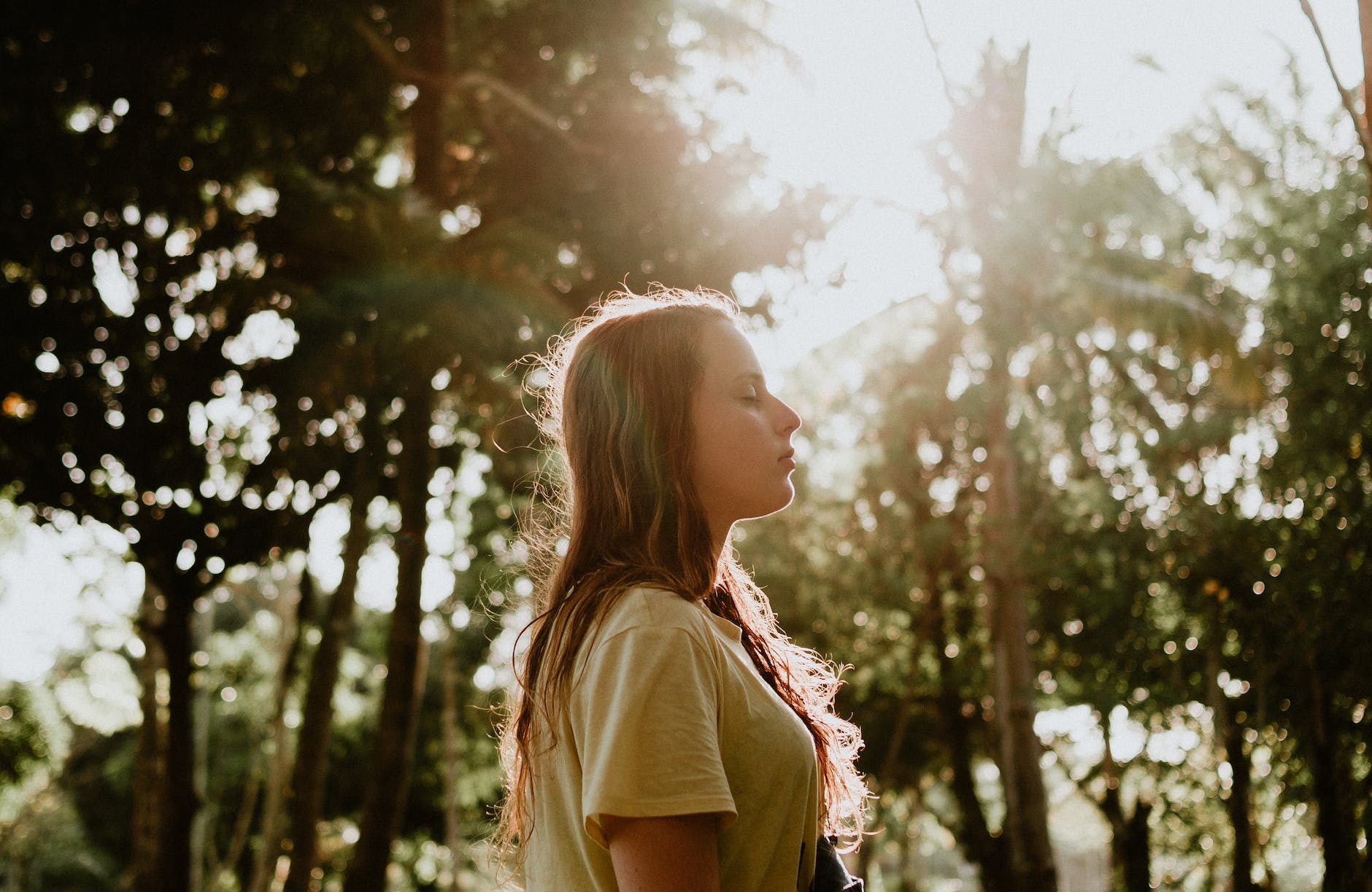 side view portrait photo of woman in yellow t shirt standing with her eyes closed with trees in the background. Thinking about the positive what ifs. 