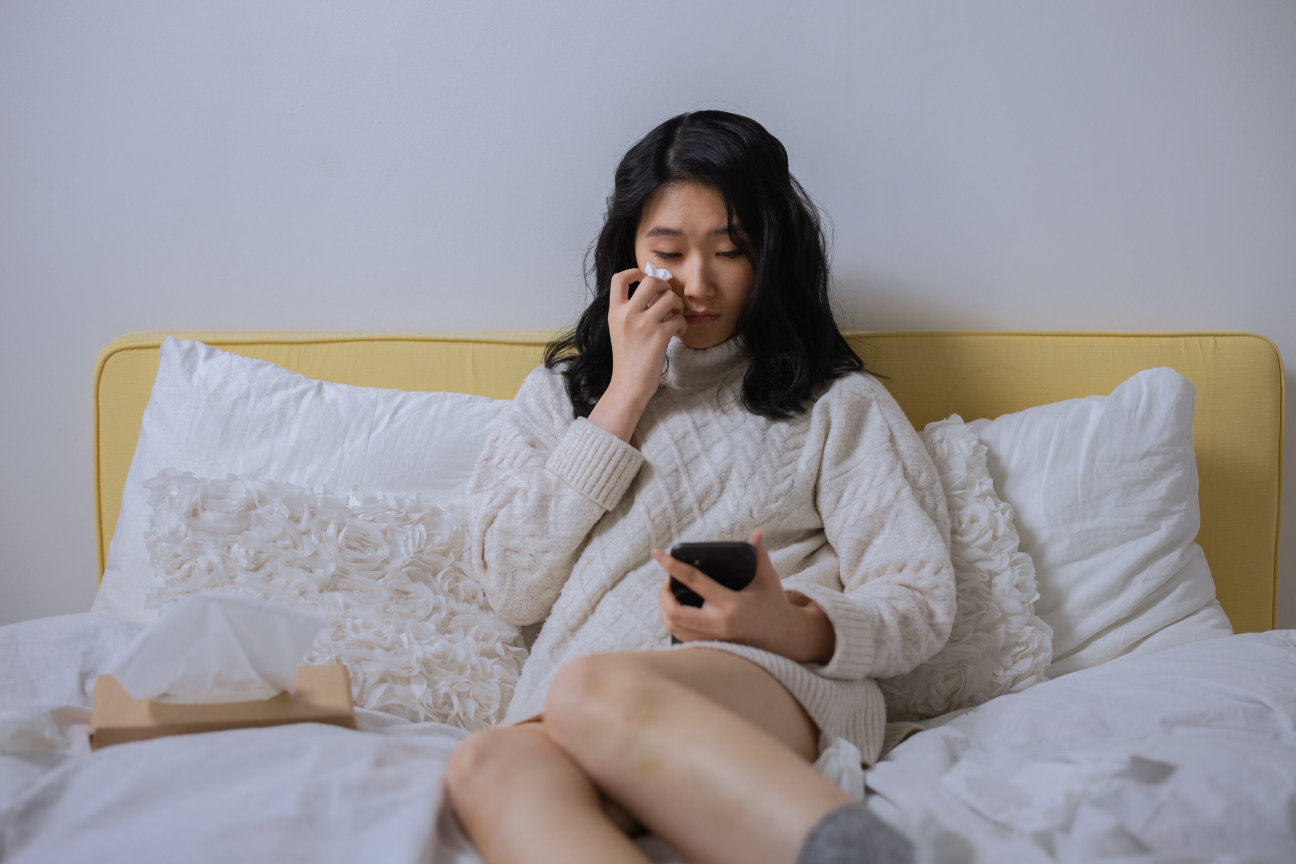 Woman sat on her bed looking at her phone with a tissue as she cries due to the negative effects social media has had on triggering her body image