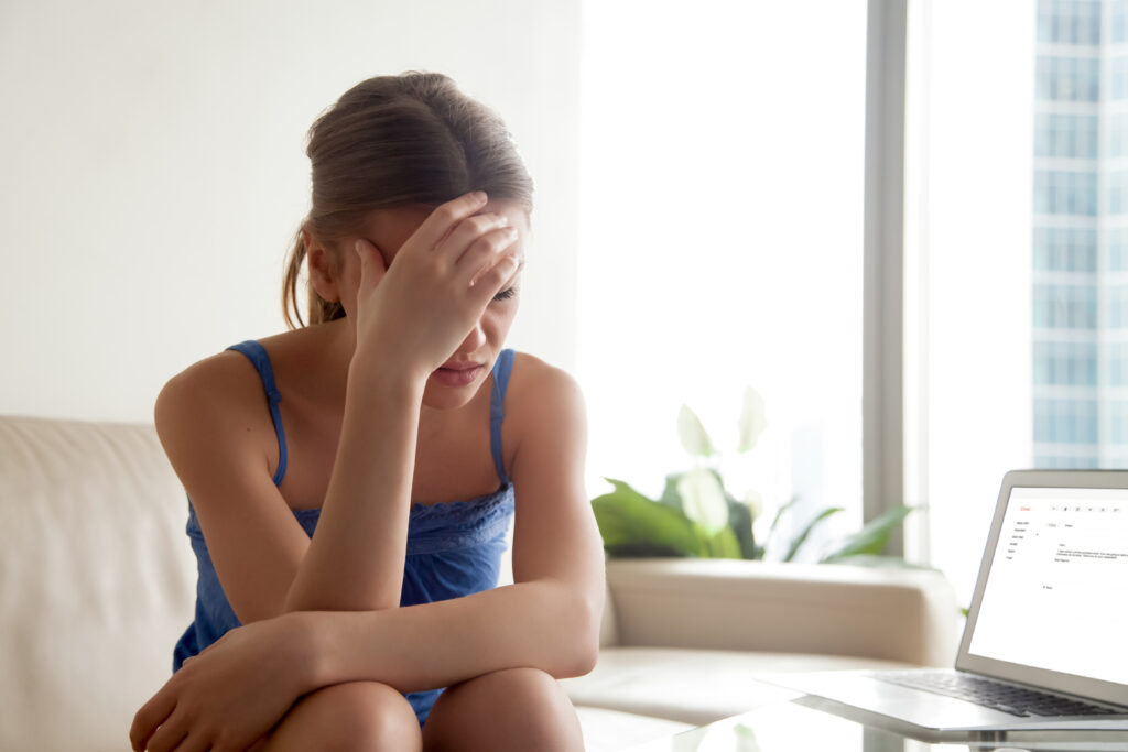 Stressed young woman in casual clothes sitting upset on sofa in front of laptop. Worried lady frustrated because of bad news in e-mail letter or message opened on computer screen, shocked of dismissal
