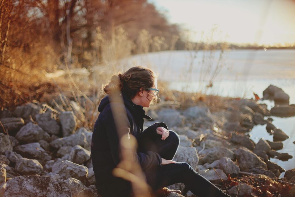 woman sitting on some stones by a lake in the winter sun as she listens to her thoughts