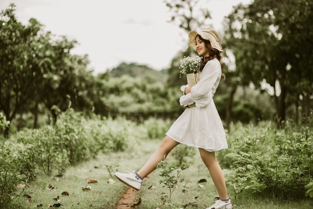 Smiling girl walking in nature holding flowers wearing white dress, white trainers and floppy sunhat. Portraying happy thoughts.