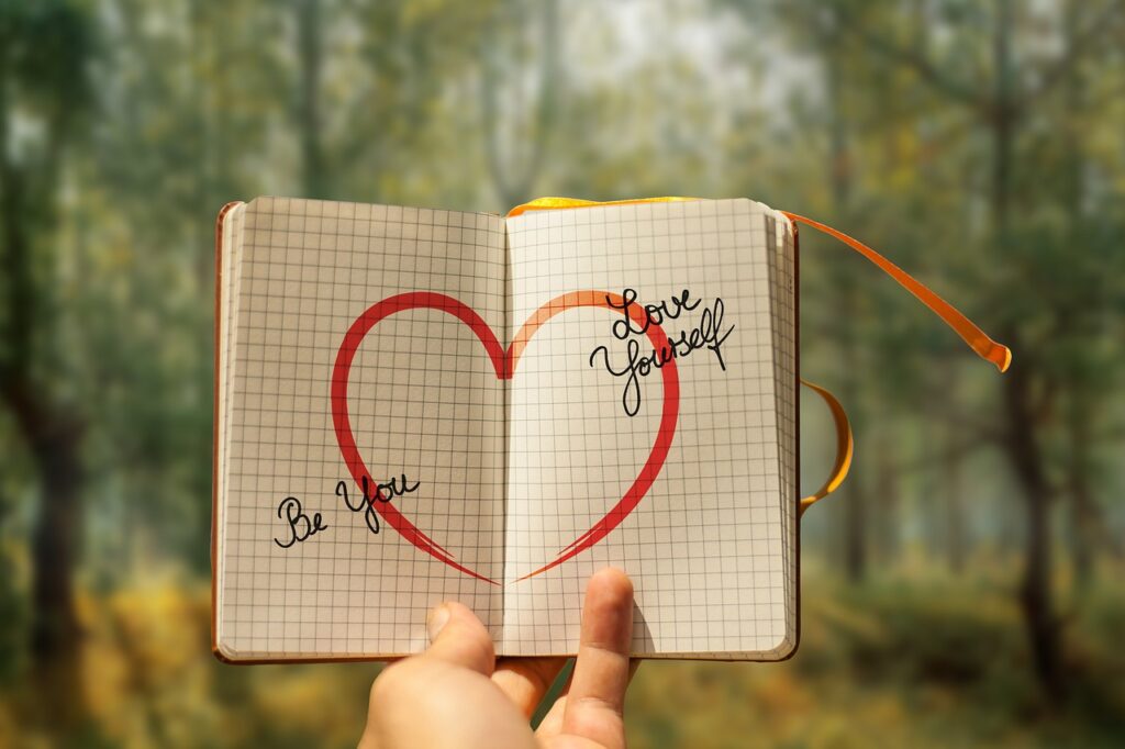open grid-lined notebook with handwritten self-love phrase 'be you love yourself' and a hand-drawn red heart across both pages.
