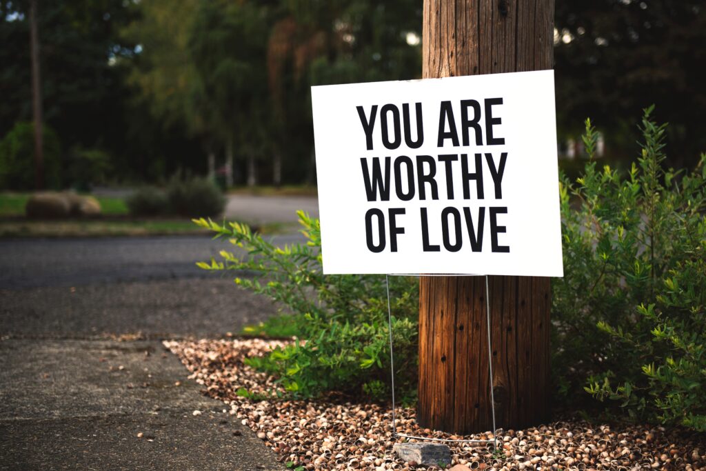 Outdoor sign in front of a wooden pillar and bush, quoting 'you are worthy of love'