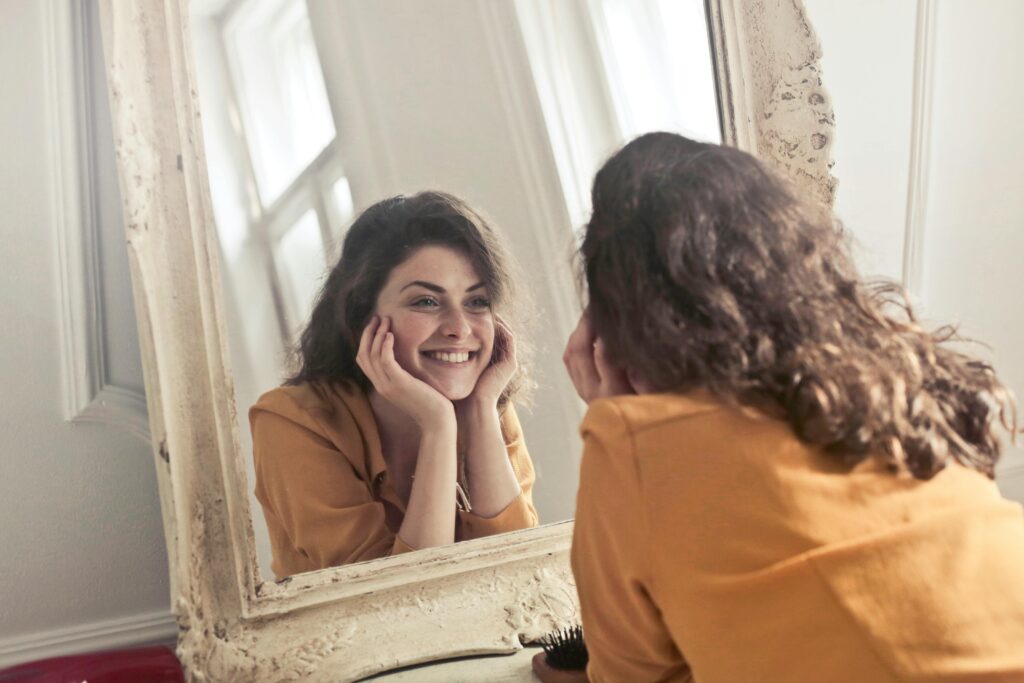 Happy smiling woman facing her reflection in mirror to support the benefits of using a daily gratitude list 