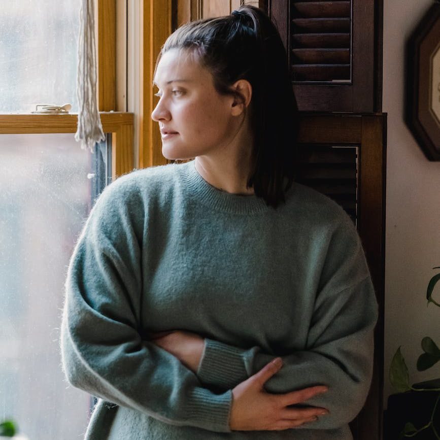 young woman in warm sweater looking outside, her facial expression and body language portraying uncertainty and insecurity.