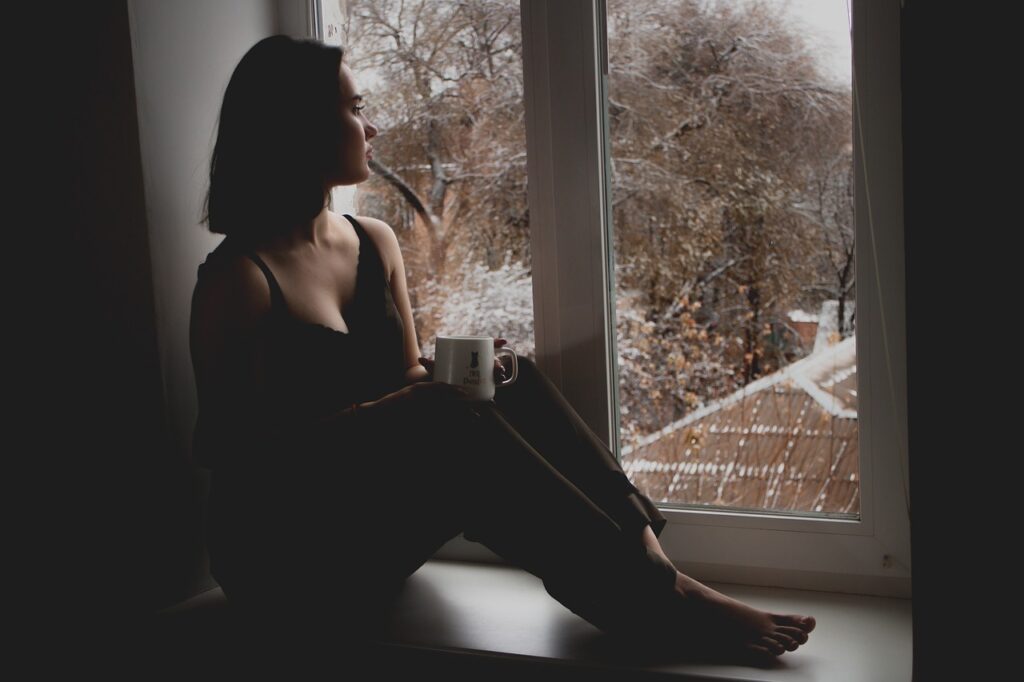 Woman sitting on window ledge with a mug, gazing out of the window. Picture used for purpose of body image quotes post, and how we all want to feel loved. 