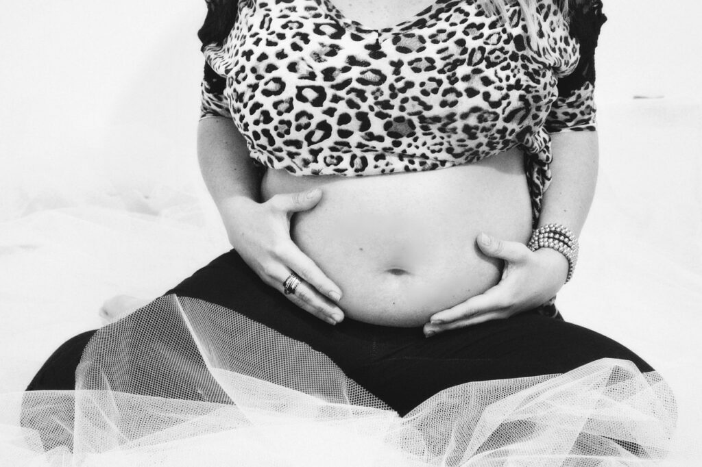 Black and white image of a pregnant woman woman wearing a leopard-print top and leggings with her bare stomach on display, sitting cross-legged holding her belly. Purpose of image to promote body appreciation.